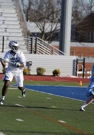 Two men playing lacrosse on a field with bleachers in the background.